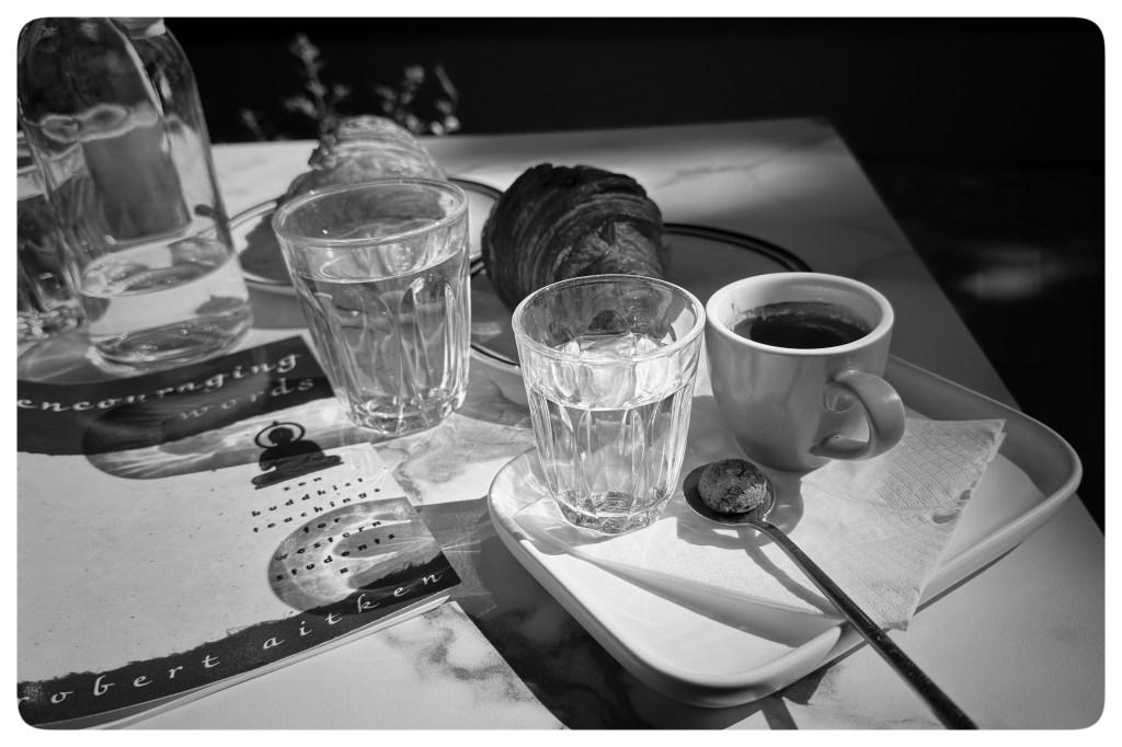 Monochrome photo of a coffee cup and croissant on a table. There are also two glasses of water and a book.