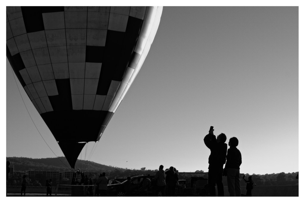 Monochrome photo of two people in silhouette taking a photo of a large hot air balloon.
