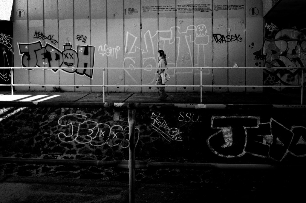 Monochrome photo of girl walking past a wall of graffitti.
