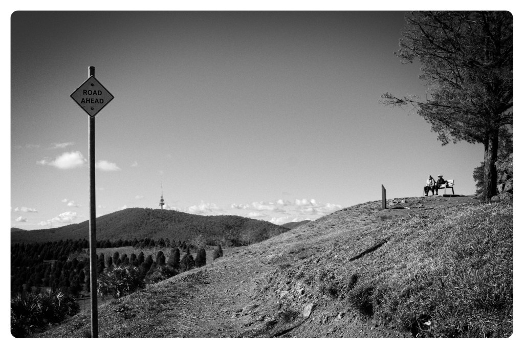 Monochrome photo of a gravel path on the side of a hill. In the distance two women are sitting on a bench. There is a sign in the foreground saying road ahead.