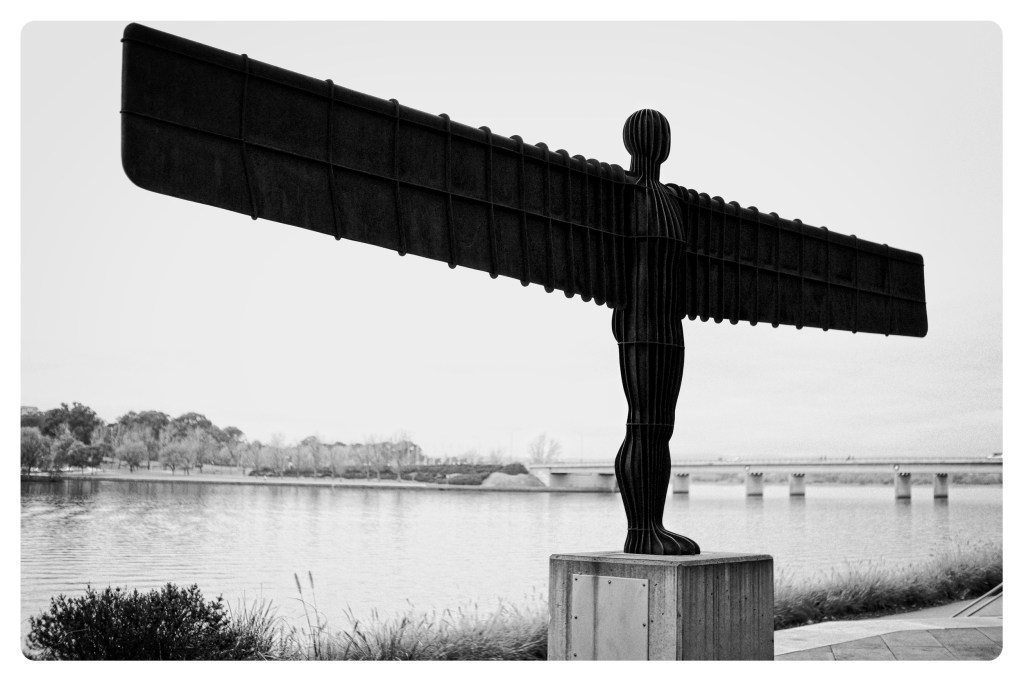 Monochrome photo of sculpture of a man with glider-like wings. In the background can be seen a lake and a distant bridge.