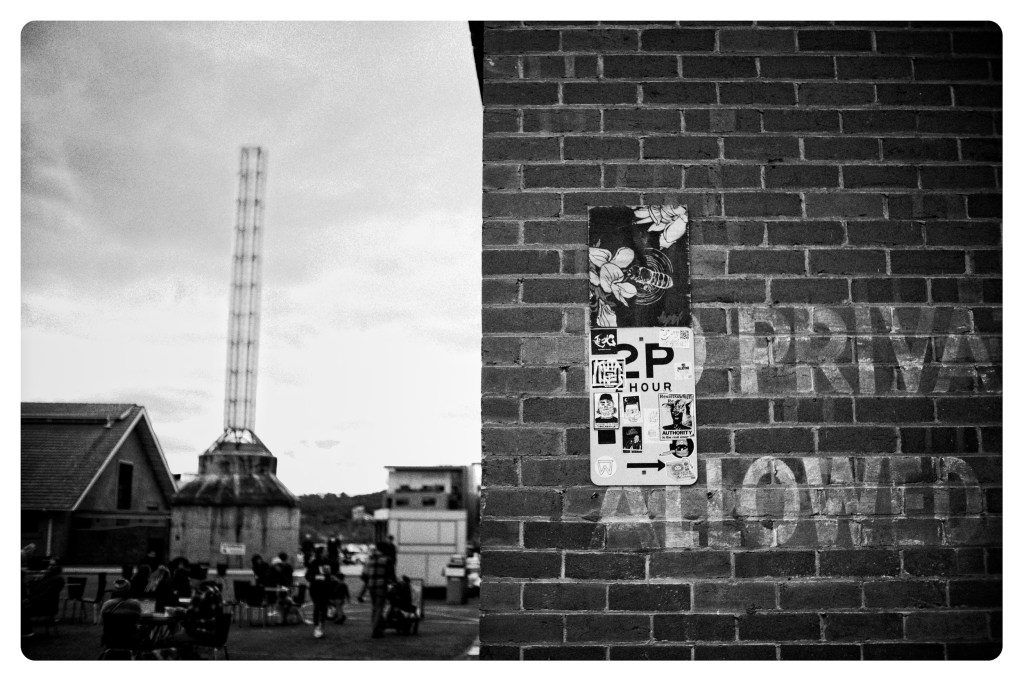 Monochrome photo of brick wall with parking sign and stickers all over it. In the background to the left of the wall can be seen people sitting at cafe tables.