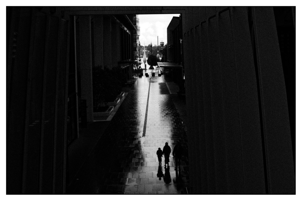 Monochrome high contrast image of two people walking in the light between two tall buildings.