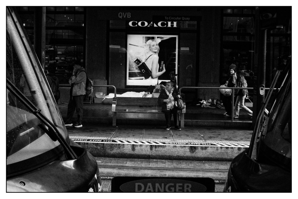 Monochrome photo of a city street scene. There is an advertising billboard on a store window depicting an expensively dressed woman. In front of it a homeless person is sleeping.