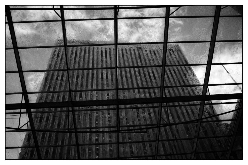 Monochrome photo of a large office building seen from below through a skylight. The sky behind is dark and wild.