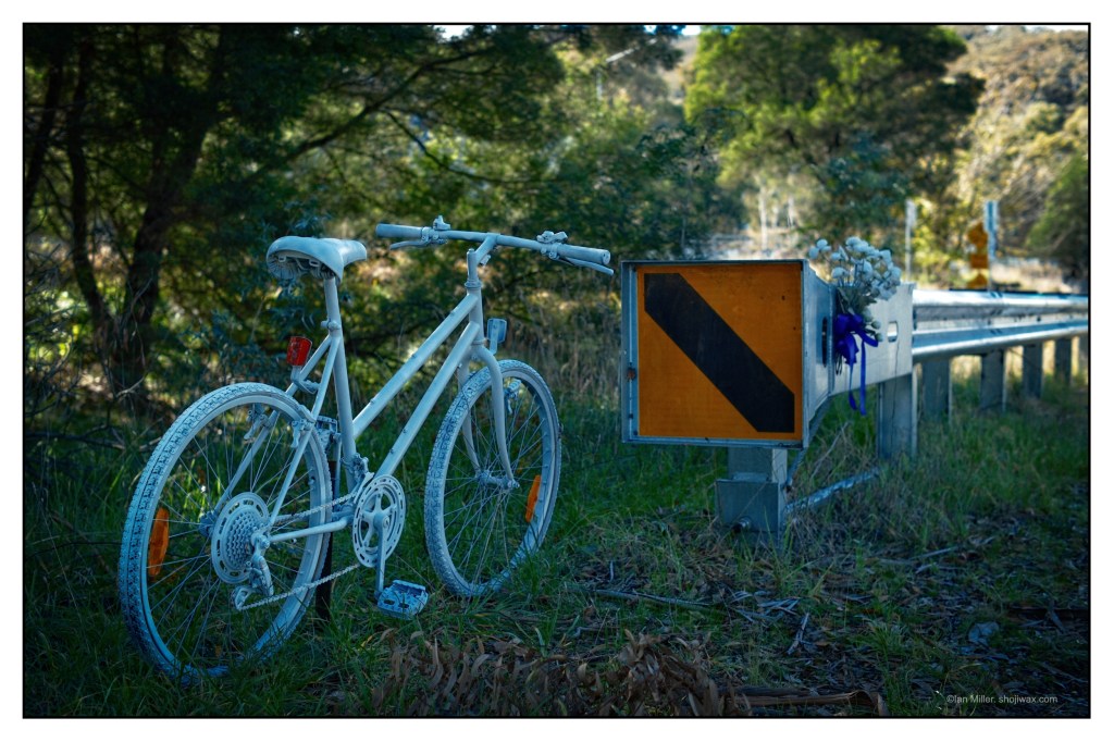 Colour photo of a 'ghost bike', painted white and located next to a road. In the background can be seen a floral tribute.