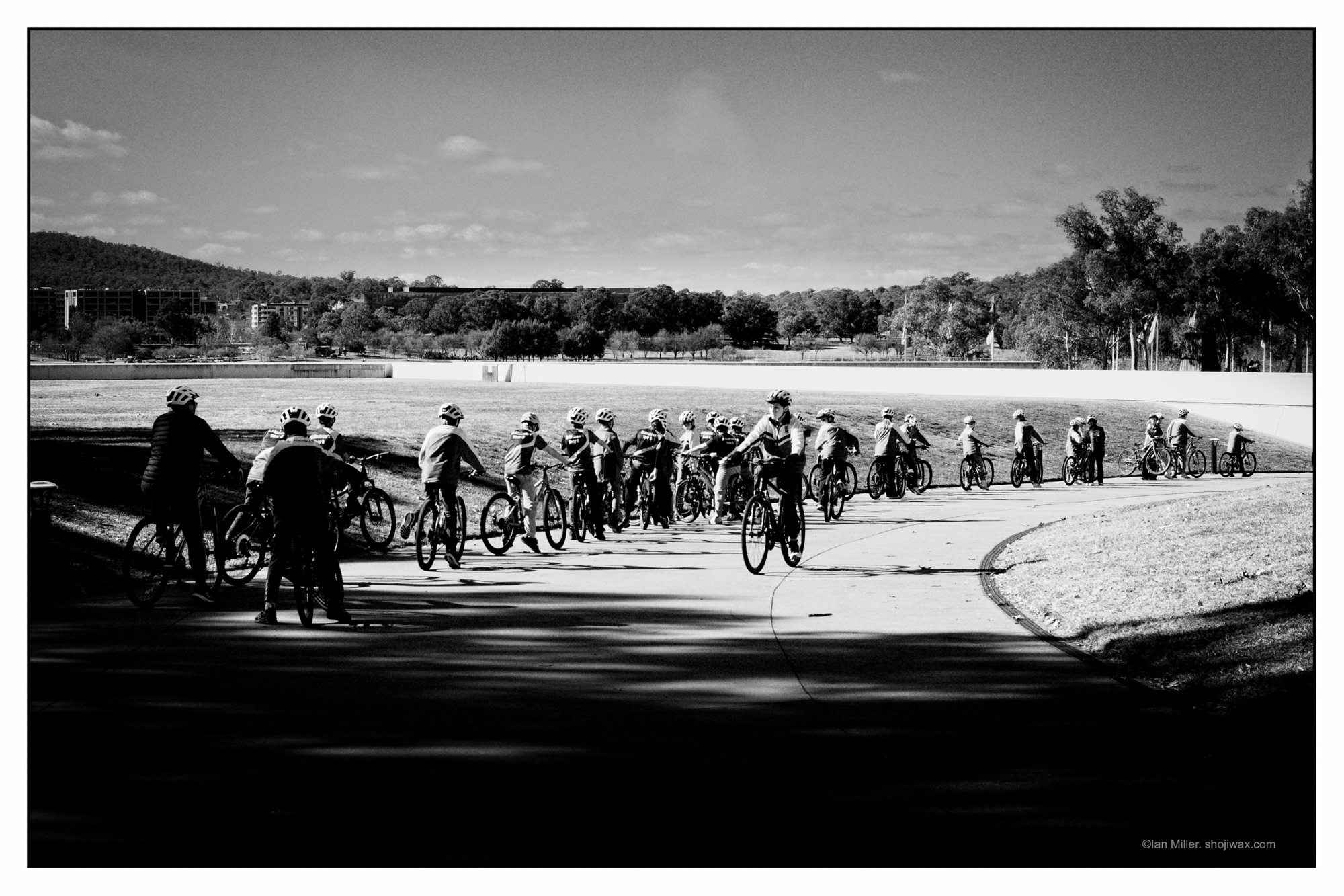 Monochrome photo of a long line of school kids riding their bikes down a wide sweeping concrete path.