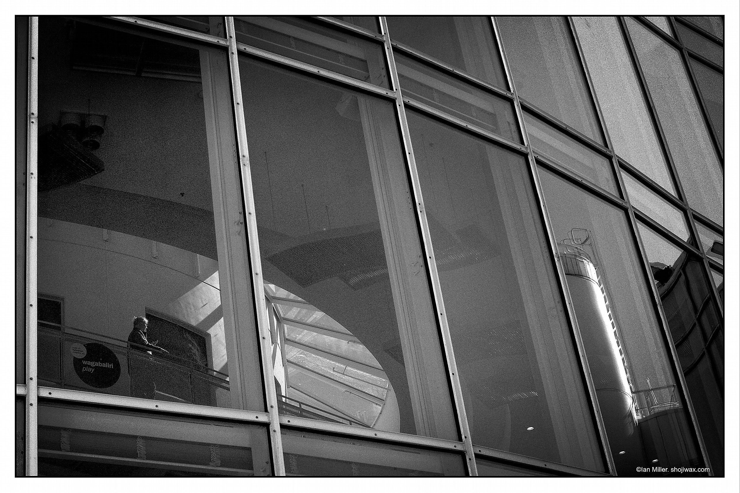 Monochrome photo of a man looking down at his phone seen through a large panel window. He is lit by a pool of light amongst the complex external reflections.