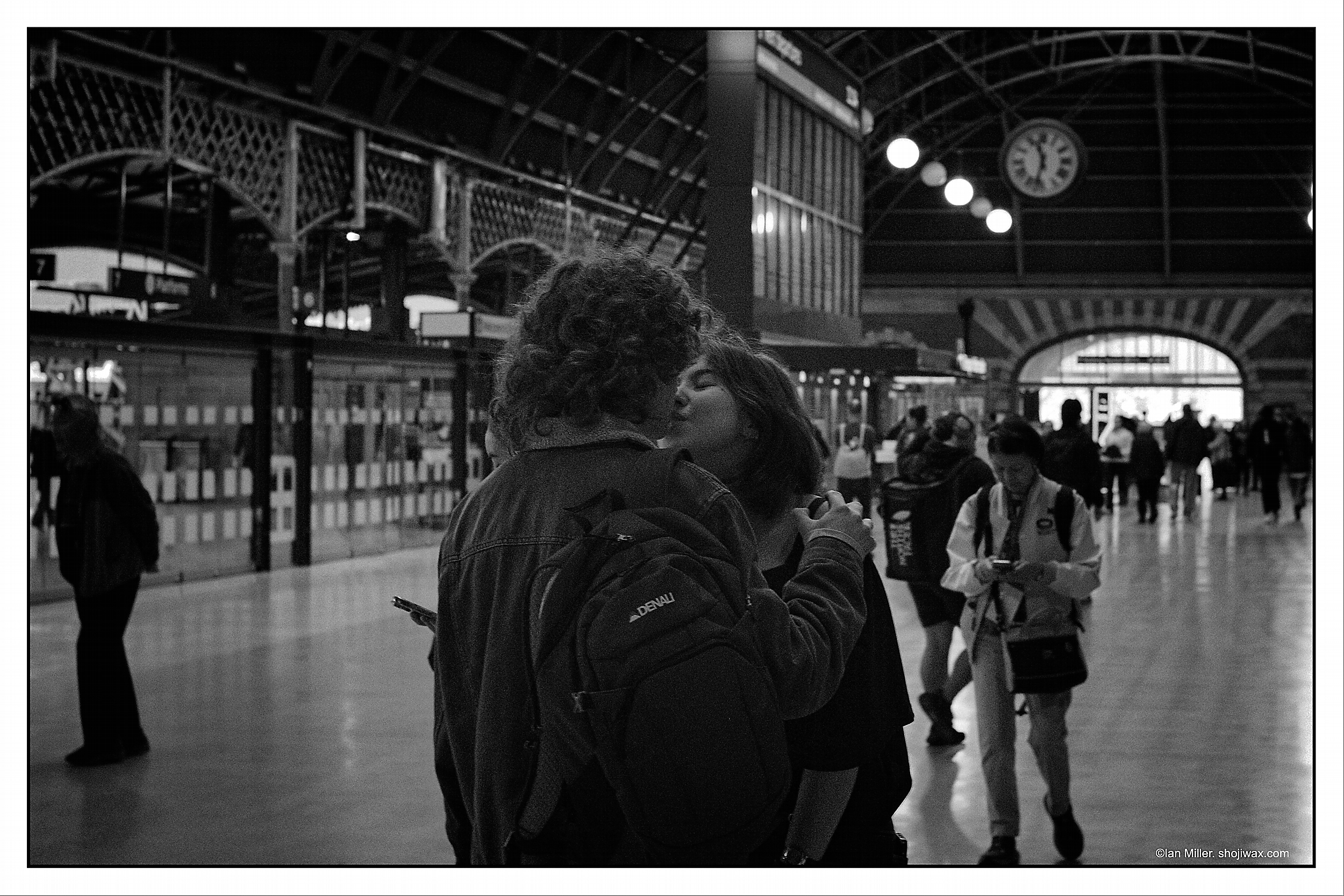 Monochrome photo of a young couple kissing in the middle of a train station.