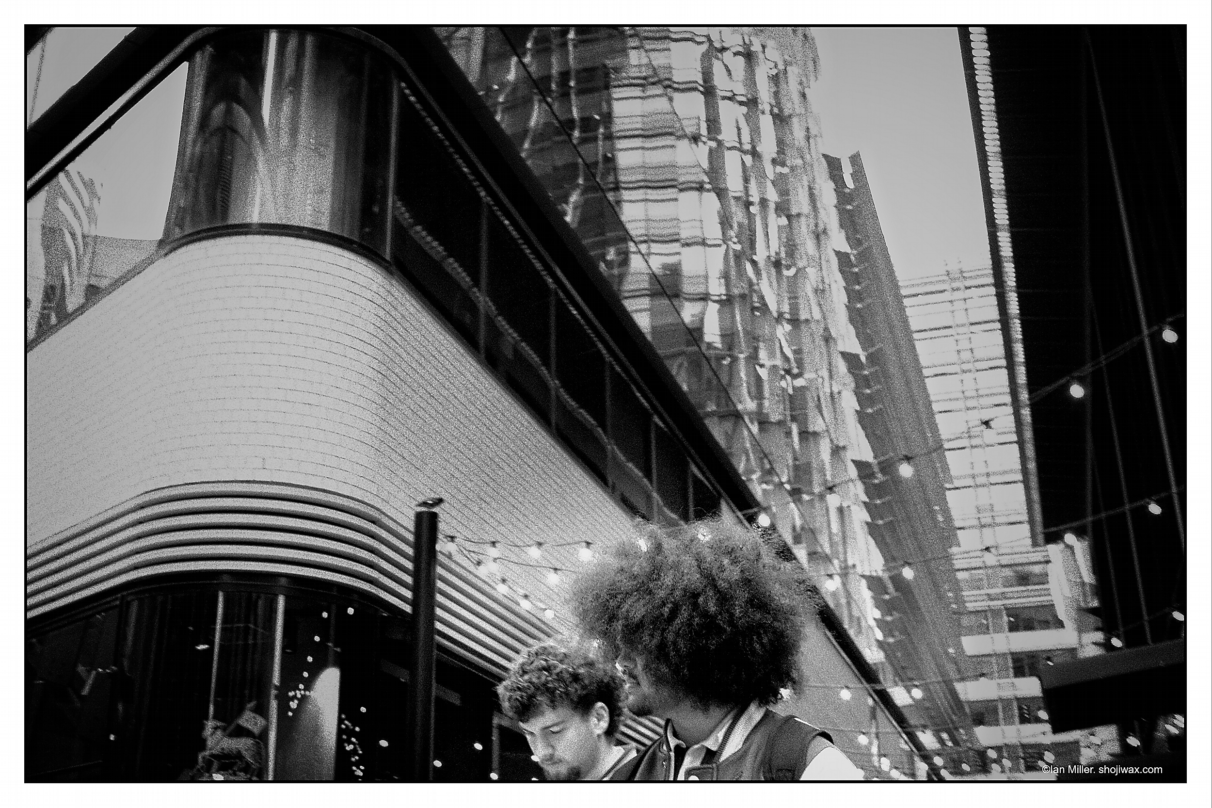 Monochrome photo of two young men. One has a large afro hairstyle. They are seen at the bottom of the image. Behind them are large buildings.