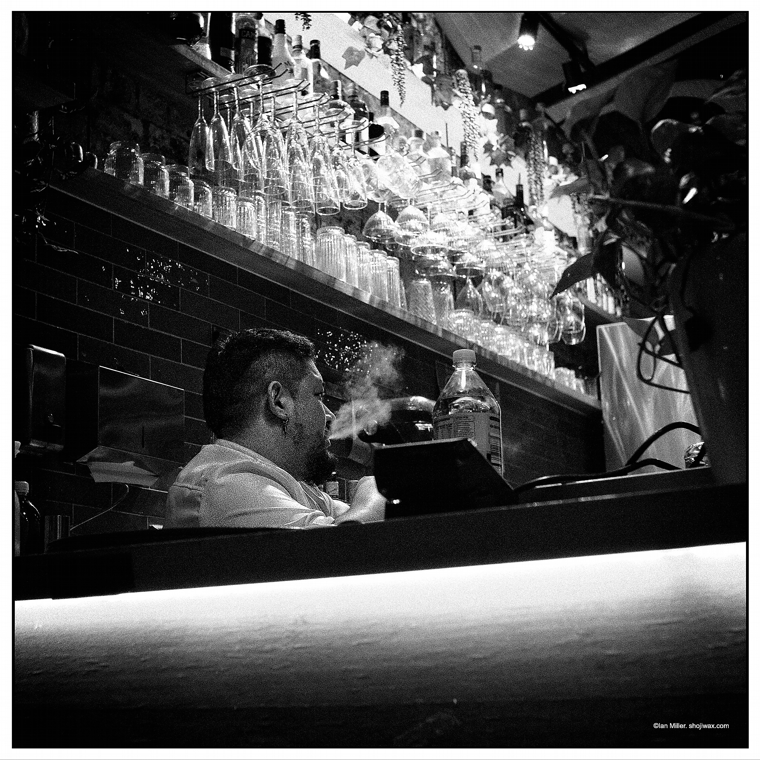 Monochrome photo of a man sitting at a bar exhaling on a vape.