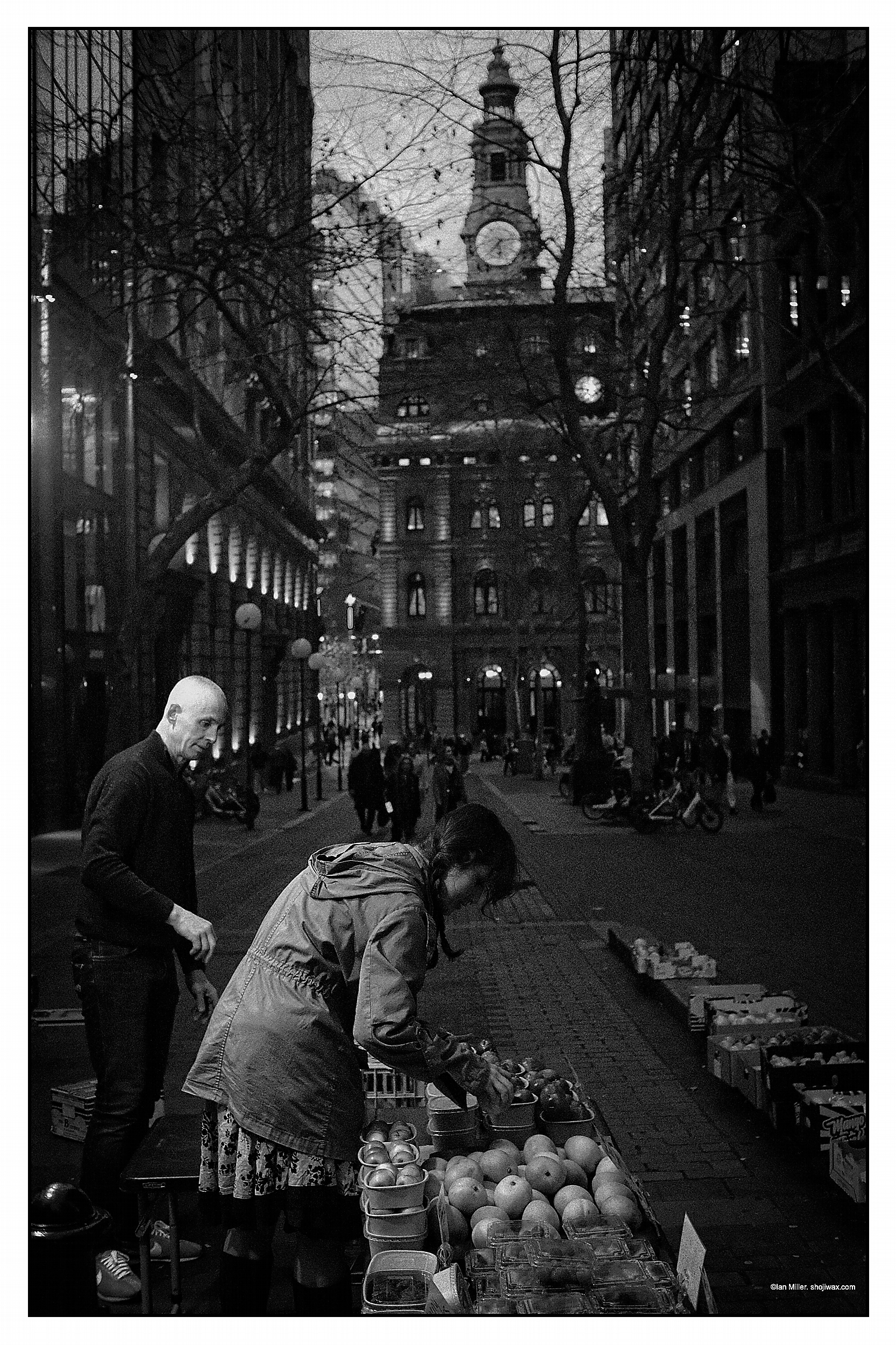 Monochrome photo taken at night of a woman bending over to pick oranges at a street vendor fruitstand.