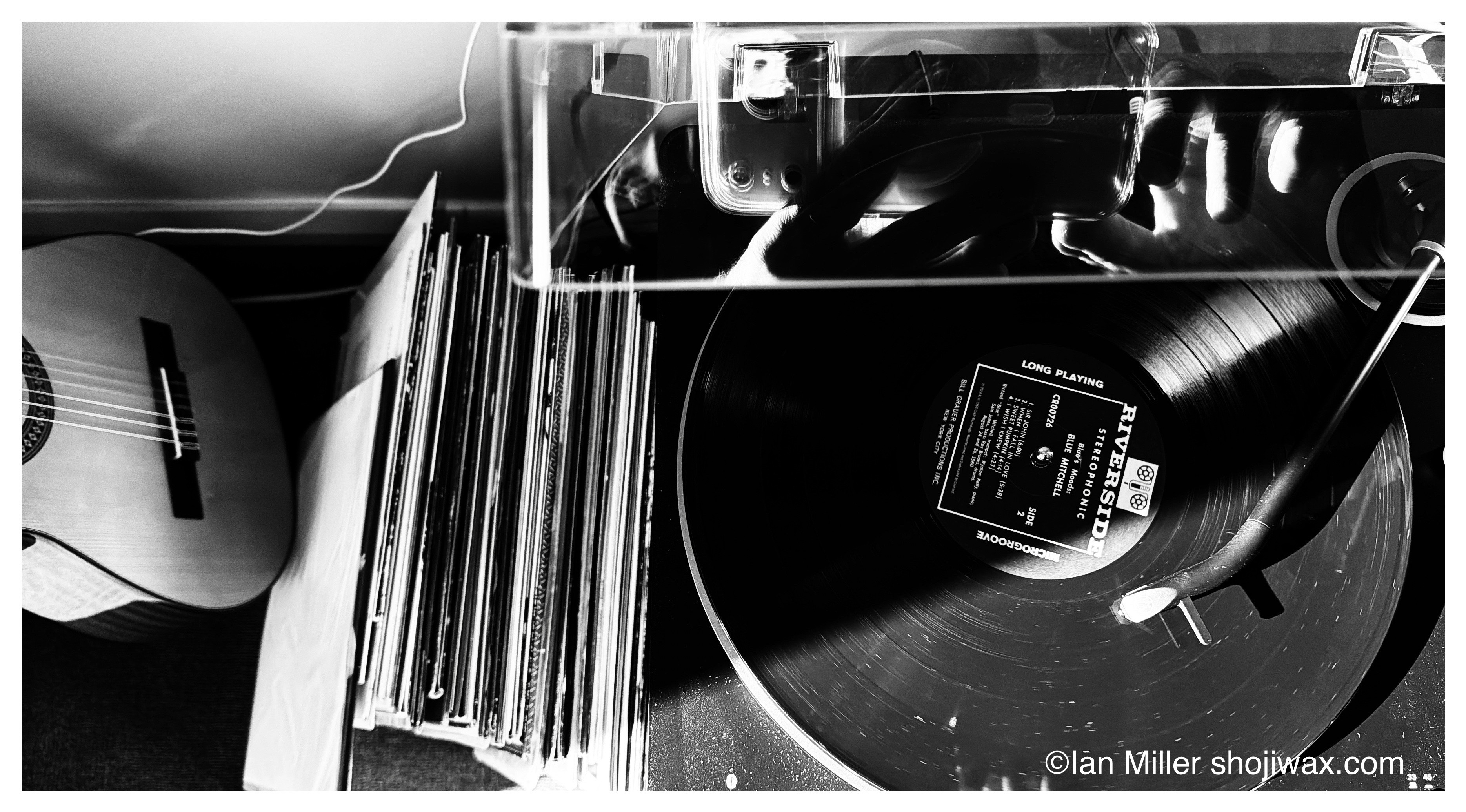 Monochrome photo of an LP record player from above. Besides the player is a stack of records and a classical guitar.