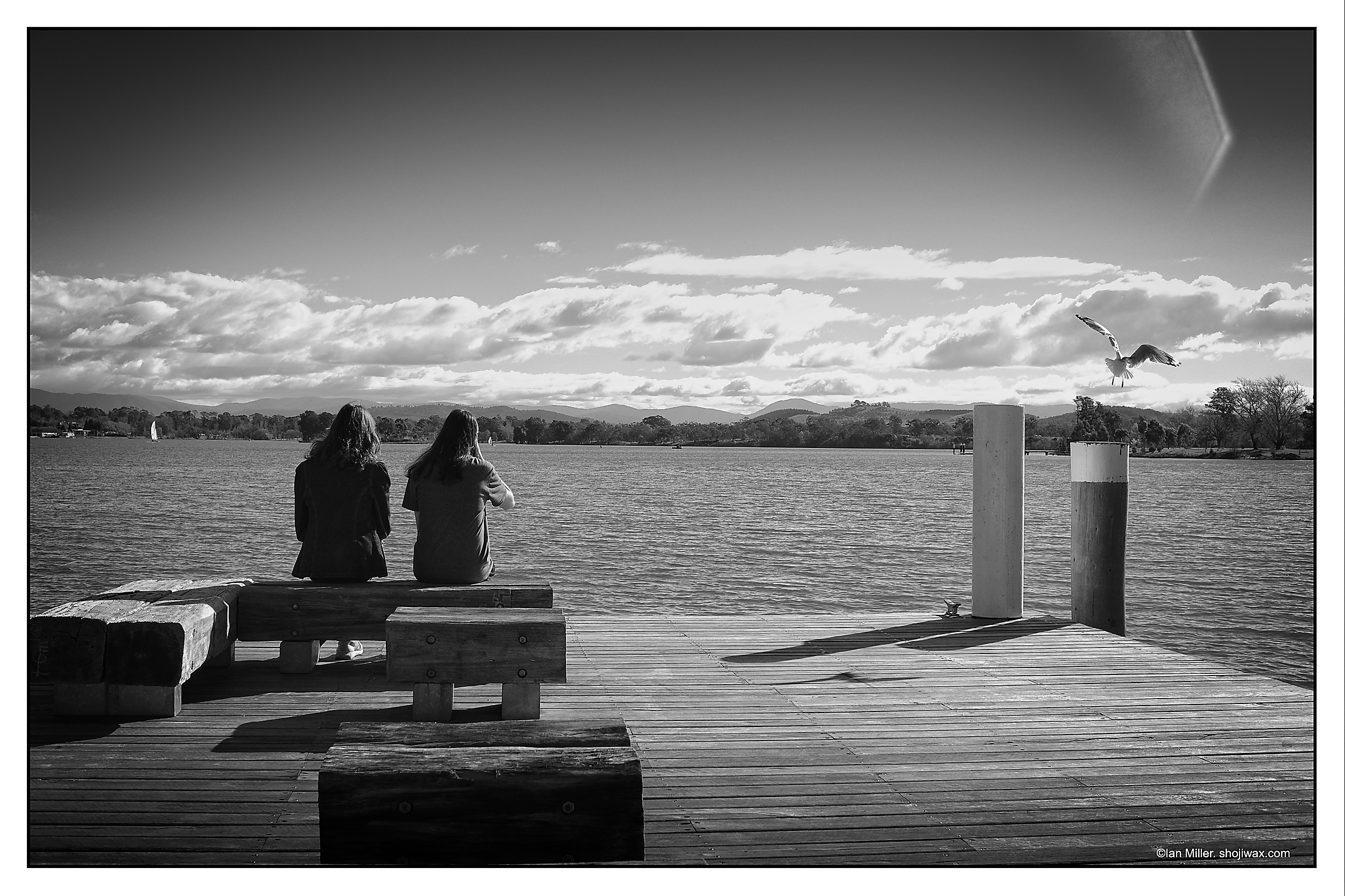 Monochrome photo of two people sitting on the edge of a lake jetty. To the left a seagull is flying way. In the distance can be seen mountain ranges.