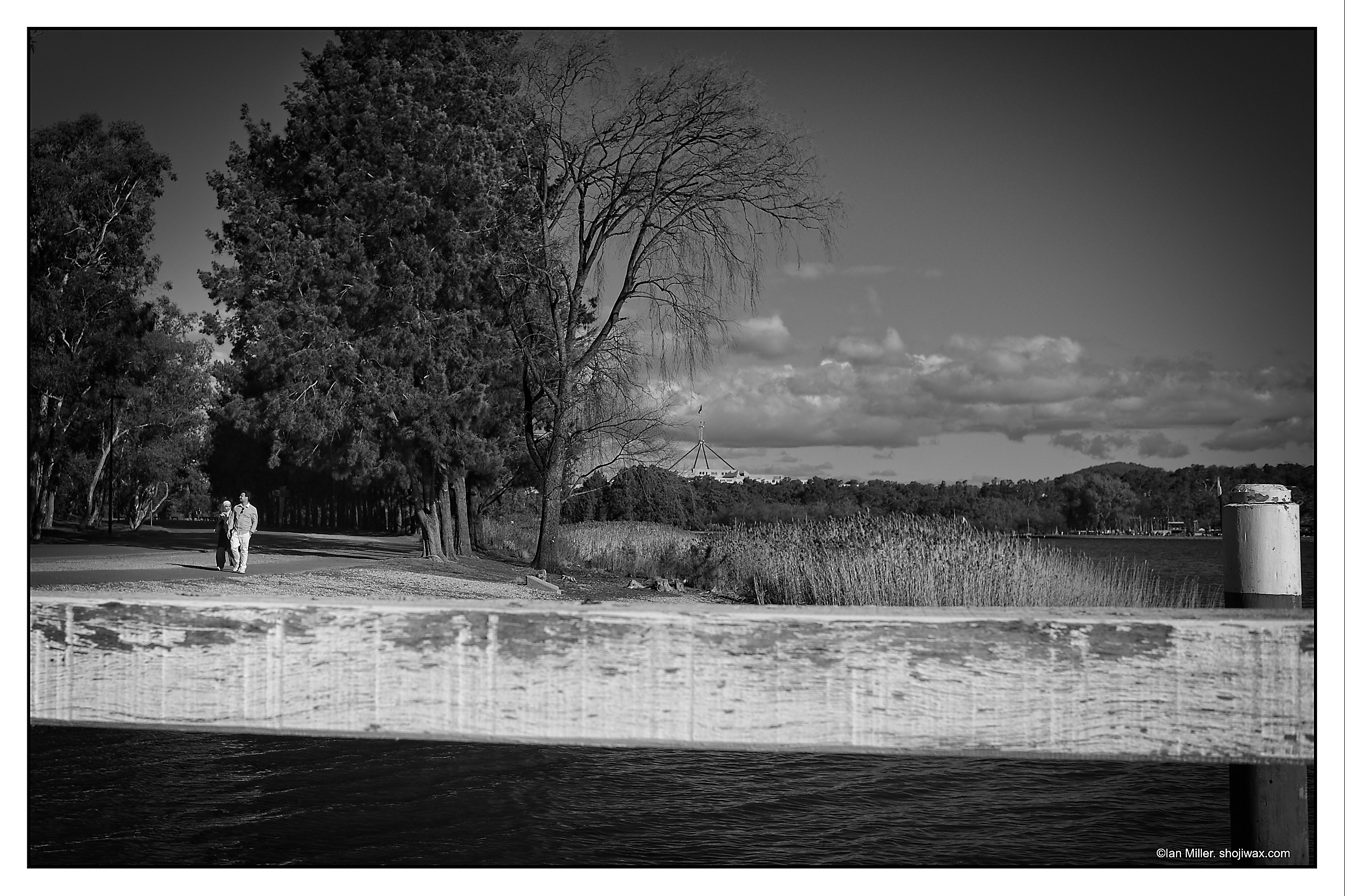 Monochrome photo of two people walking in the distance amongst a lake and treelined scene.